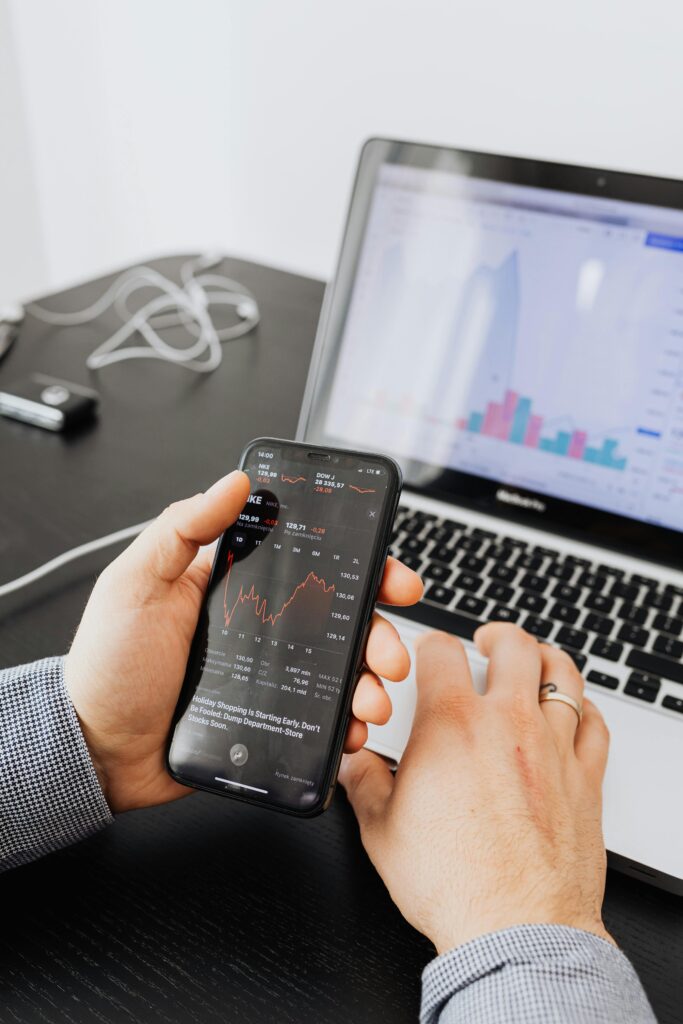 Hands holding smartphone analyzing stock market data next to laptop displaying charts.